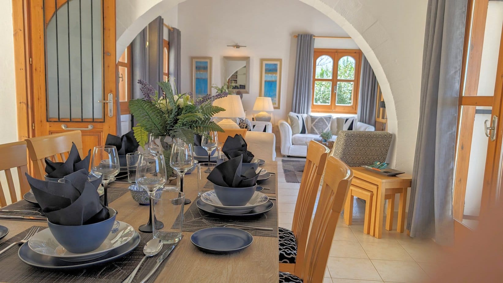 The dining table at Villa Koumos is set for a meal and beyond the table is a view of the lounge with comfortable seating